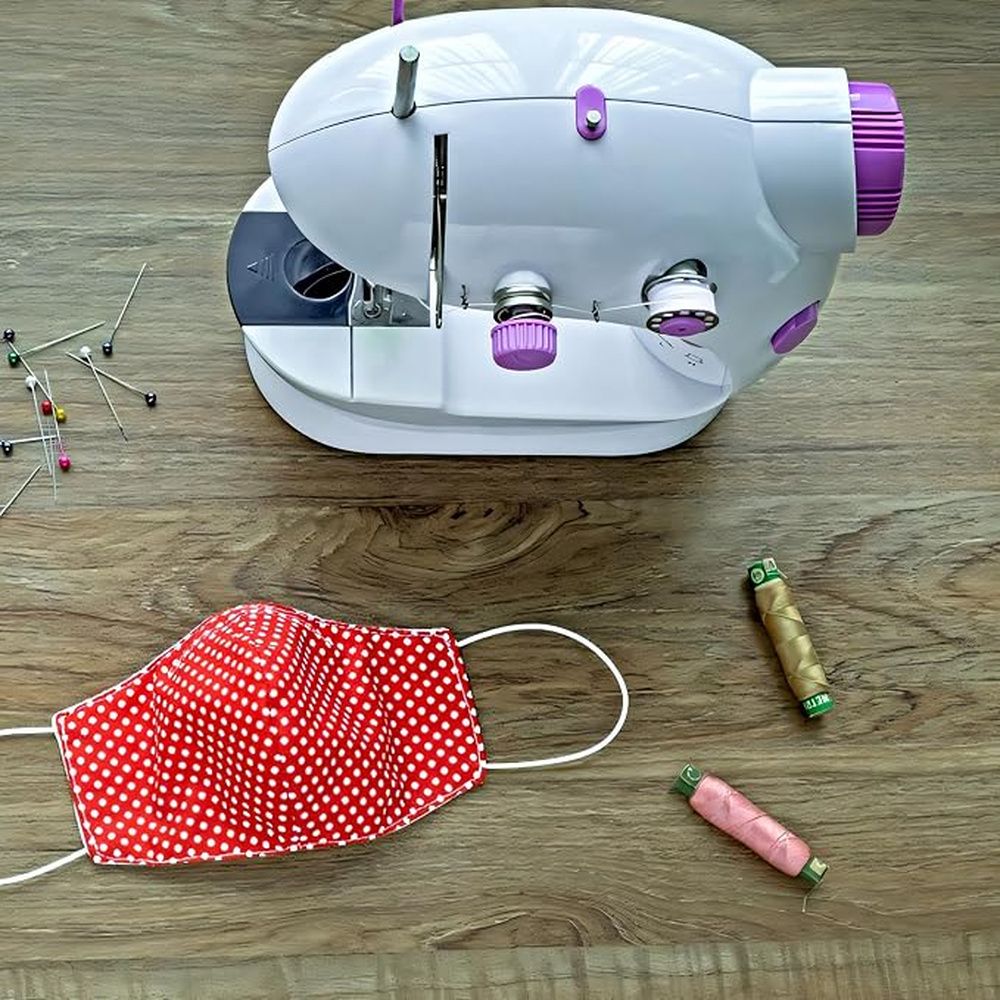 Máquina de coser portátil blanca y violeta junto a una mascarilla de tela roja, hilos y alfileres sobre una mesa de madera.