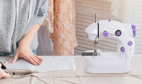 Mujer preparando una tela sobre una mesa junto a una máquina de coser portátil blanca con controles violetas.