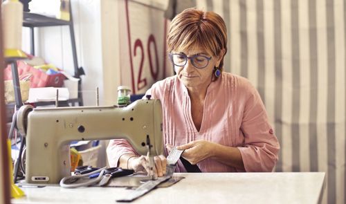 Mujer con gafas cosiendo una tela a máquina en un taller, con tijeras sobre la mesa.