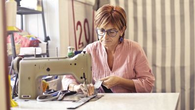 Mujer con gafas cosiendo una tela a máquina en un taller, con tijeras sobre la mesa.