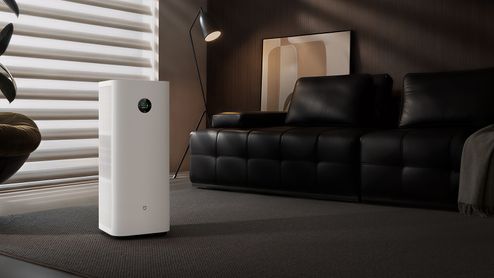 Mijia Smart Air Purifier 6 Plus blanco junto a una ventana y un sofá negro en un salón.