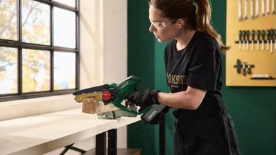 Una mujer con gafas y guantes de protección corta una pieza de madera con una minisierra Parkside en un taller.