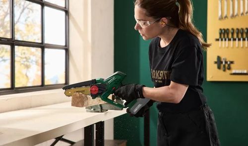 Una mujer con gafas y guantes de protección corta una pieza de madera con una minisierra Parkside en un taller.