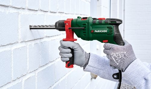 Taladro de impacto Parkside verde y rojo perforando una pared de ladrillo blanco, sujetado con guantes de trabajo.