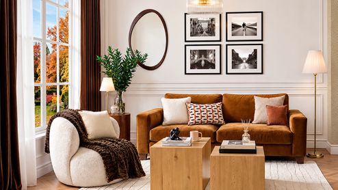 Salón con sofá marrón, sillón blanco con manta, mesas de madera y cojines en tonos beige y teja.