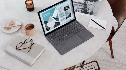 Ordenador portátil sobre una mesa junto a una taza de café, un cuaderno y material de trabajo.