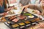Parrilla raclette sobre una mesa con verduras, carne y pequeñas bandejas para fundir queso durante una cena.