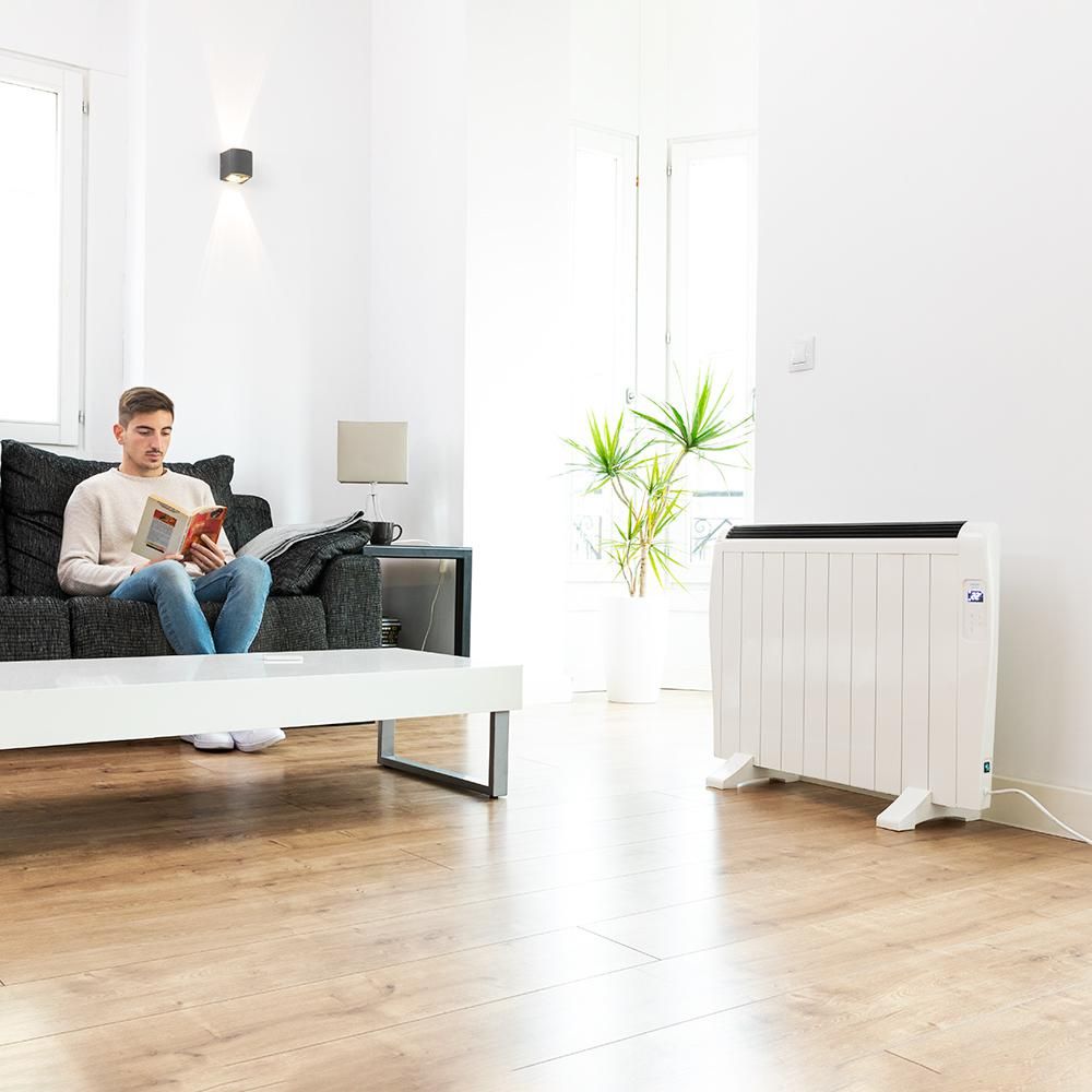 Emisor térmico Cecotec blanco sobre patas en un salón luminoso, con un hombre leyendo en el sofá.