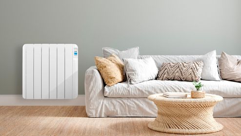 Radiador eléctrico HJM blanco instalado en una pared gris, junto a un sofá claro y una mesa de fibras naturales.