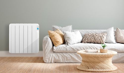 Radiador eléctrico HJM blanco instalado en una pared gris, junto a un sofá claro y una mesa de fibras naturales.