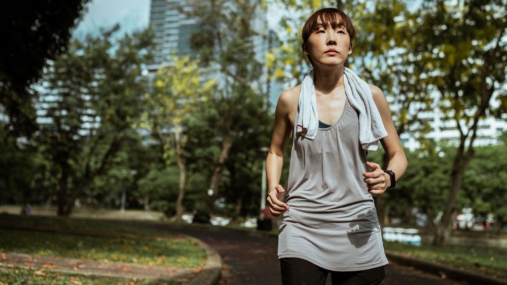 Mujer con ropa deportiva gris y una toalla al cuello corriendo por un parque.