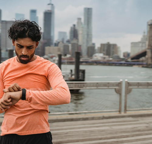 Hombre con camiseta naranja consultando su reloj deportivo junto al puente de Brooklyn.