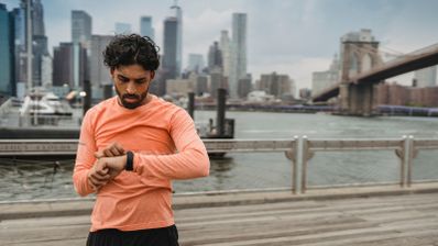 Hombre con camiseta naranja consultando su reloj deportivo junto al puente de Brooklyn.