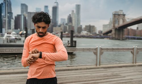 Hombre con camiseta naranja consultando su reloj deportivo junto al puente de Brooklyn.