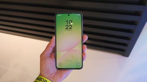Samsung Galaxy A56 sostenido en una mano, con la pantalla de bloqueo encendida y un fondo en tonos verdes y rosas.