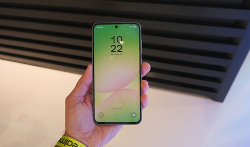 Samsung Galaxy A56 sostenido en una mano, con la pantalla de bloqueo encendida y un fondo en tonos verdes y rosas.