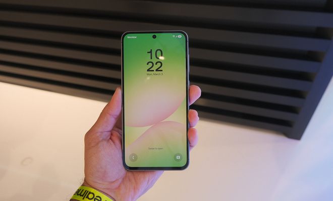 Samsung Galaxy A56 sostenido en una mano, con la pantalla de bloqueo encendida y un fondo en tonos verdes y rosas.
