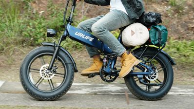 Primer plano de una bicicleta eléctrica UrLife T5 azul en marcha, con neumáticos anchos y equipaje en el portaequipajes trasero.