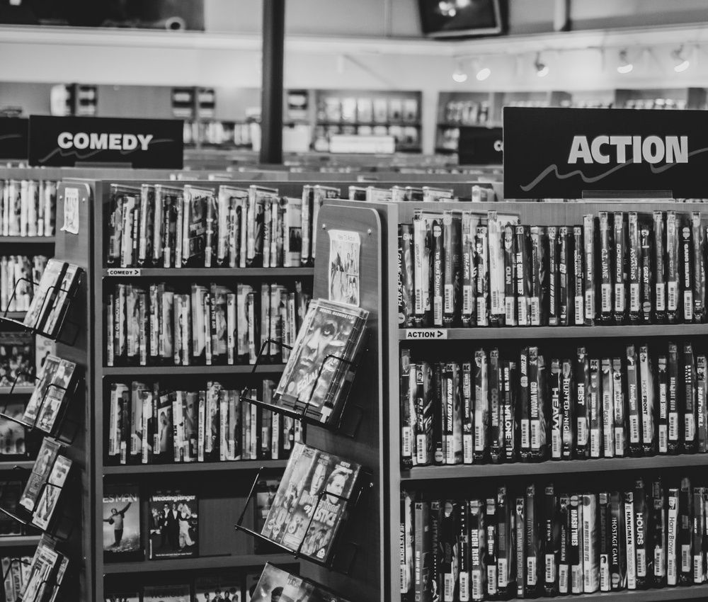 Fotografía en blanco y negro de un videoclub con estanterías de películas organizadas por géneros.