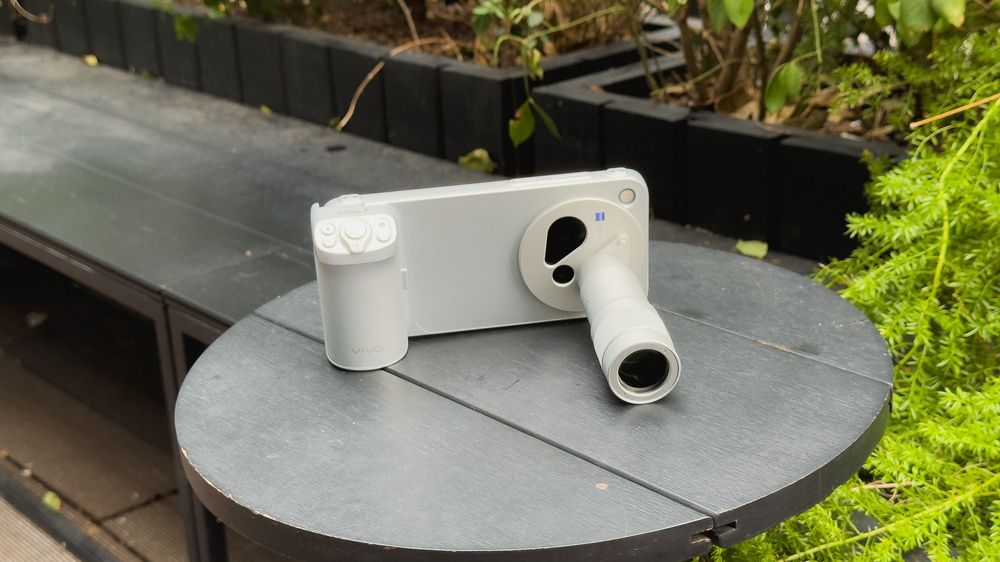 Vivo X500 Pro Max con empuñadura y objetivo externo acoplados, sobre una mesa junto a unas plantas.