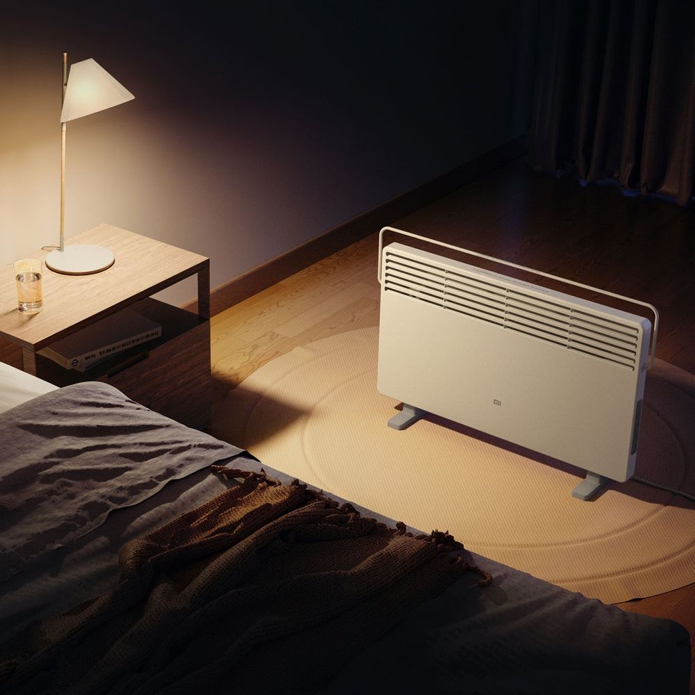 Calefactor Xiaomi Mi Smart Space Heater S en un dormitorio, junto a una cama y una mesilla con lámpara encendida.