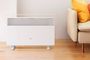 Calefactor Xiaomi Mi Smart Space Heater S blanco sobre suelo de madera, junto a un sofá con cojines amarillos y naranjas.