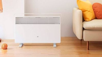 Calefactor Xiaomi Mi Smart Space Heater S blanco sobre suelo de madera, junto a un sofá con cojines amarillos y naranjas.