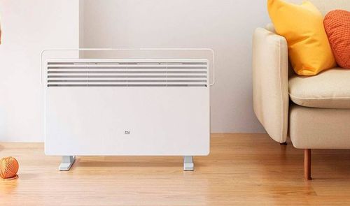 Calefactor Xiaomi Mi Smart Space Heater S blanco sobre suelo de madera, junto a un sofá con cojines amarillos y naranjas.